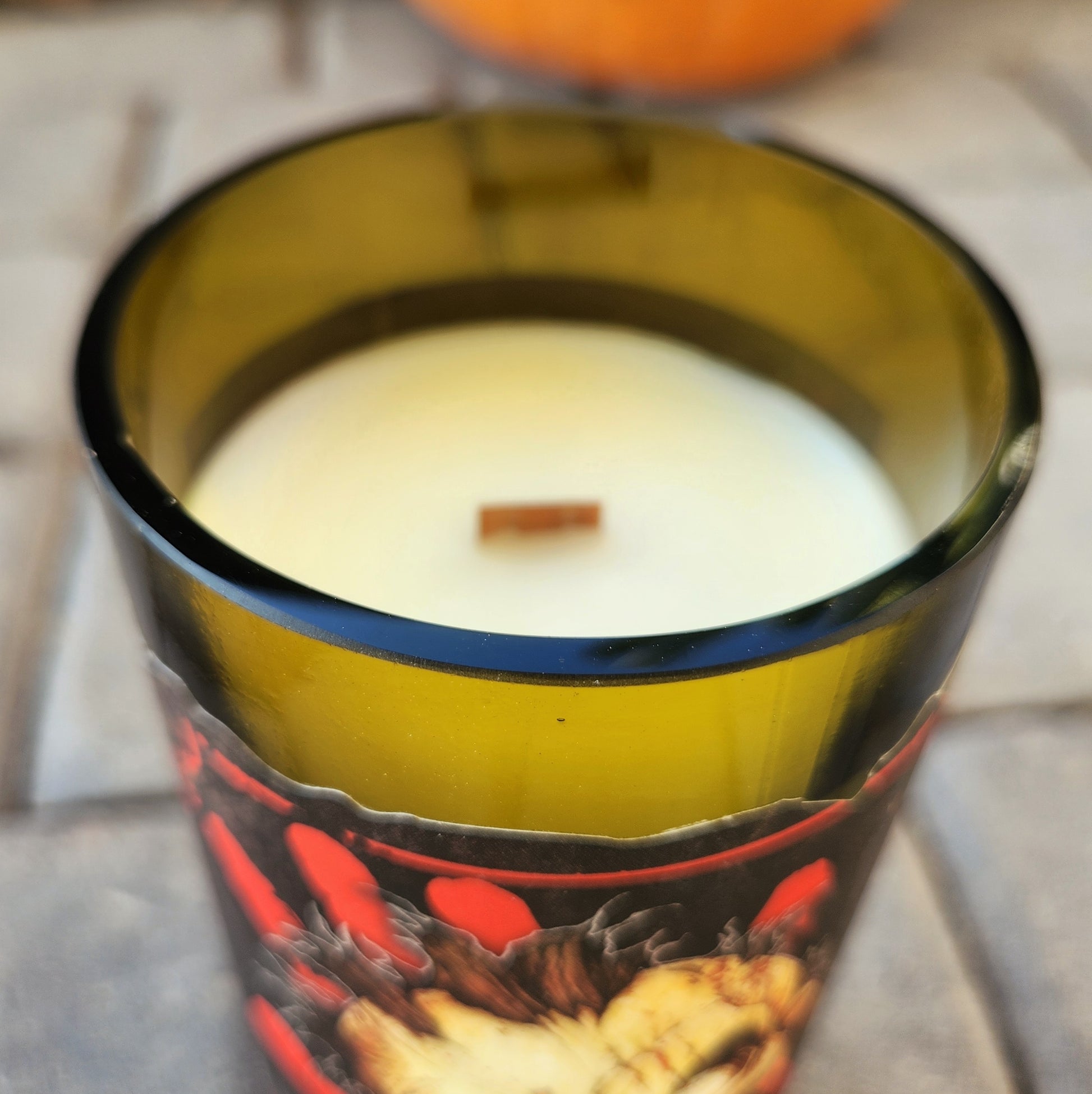 Close-up of a hand-poured eco-friendly candle in a recycled wine bottle with a shiny smooth rim. The label features a dramatic zombie illustration from Dead Zed Red wine, highlighting the non-toxic coconut soy wax and crackling wooden wick.