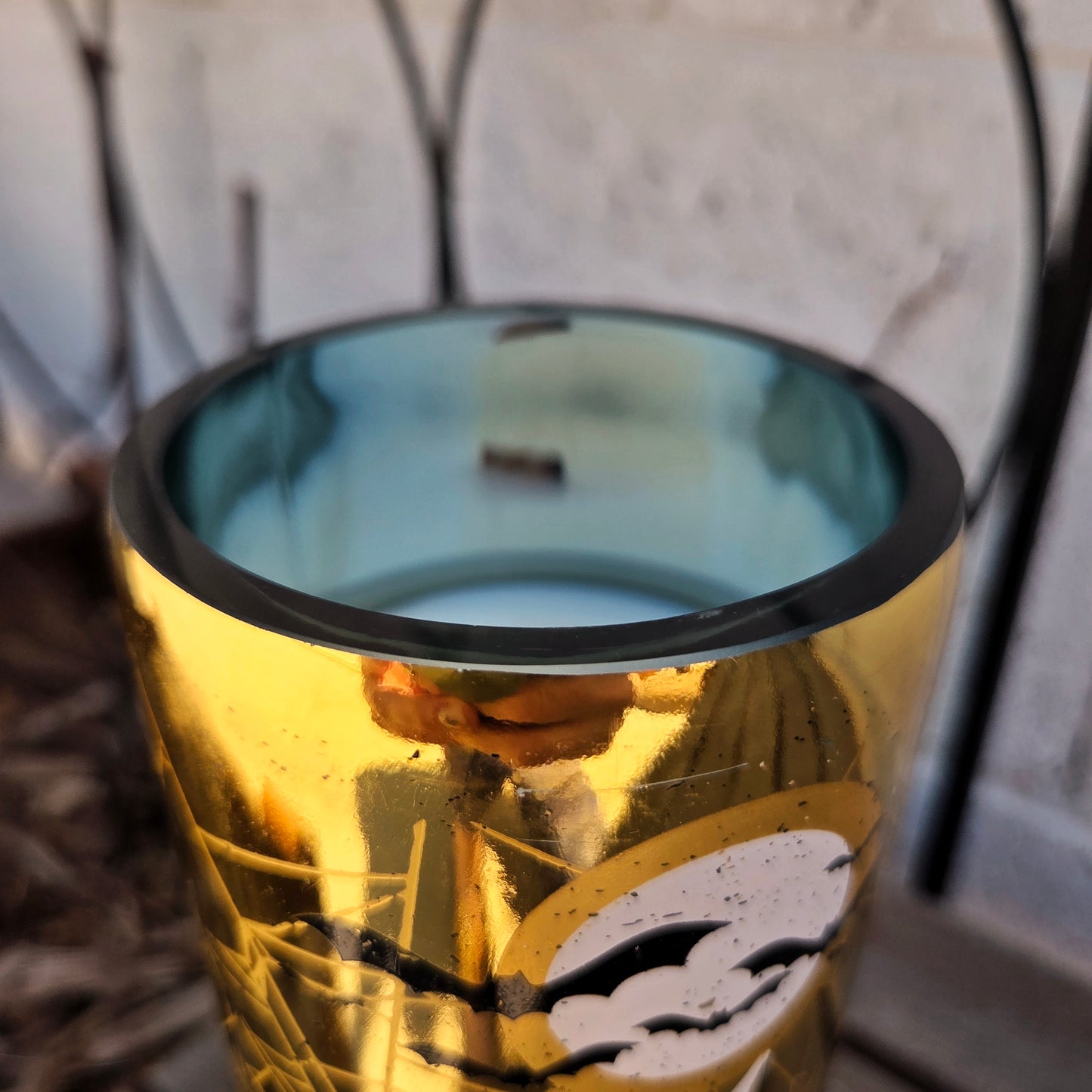 Close-up of the shiny smooth glass rim on a gold wine bottle candle, showing the reflective metallic design with ghostly Halloween artwork. The coconut soy wax and wooden wick sit neatly inside for an eco-friendly burn.
