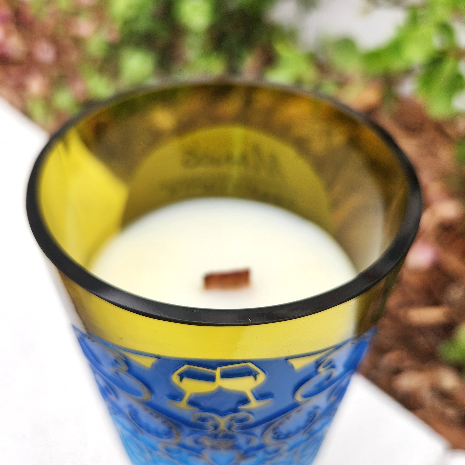 Close-up of the glossy, smooth rim of a recycled wine bottle candle with a vivid blue embossed sugar skull design. The clear glass subtly transitions into a soft yellow-green tint, housing a hand-poured coconut soy wax blend and a wooden wick for a clean, non-toxic burn.

