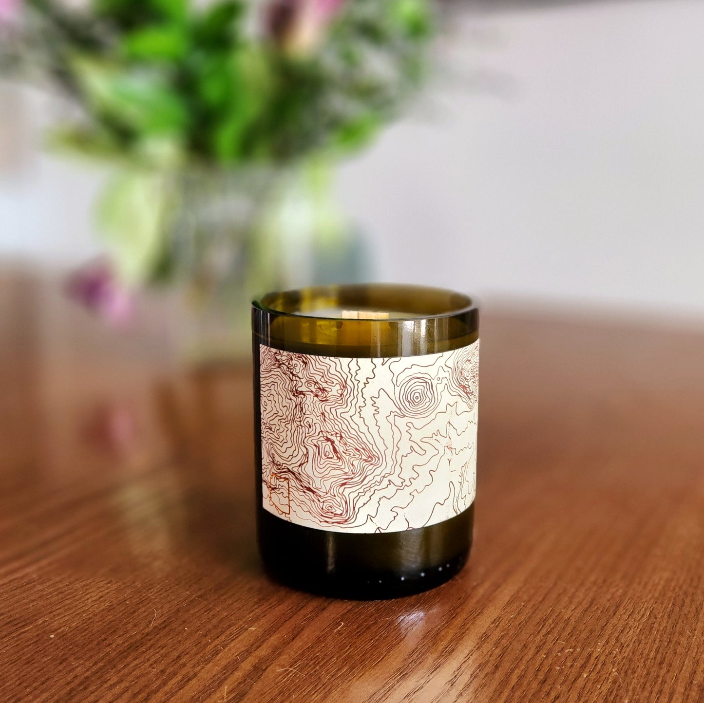 A hand-poured coconut soy wine bottle candle housed in a recycled Rubrix wine bottle. The label features an intricate topographical map design, adding an adventurous and artistic touch. This eco-friendly wooden wick candle has a polished glass rim, highlighting expert craftsmanship. Placed on a smooth wooden table with a softly blurred floral arrangement in the background, this sustainable wine bottle candle is perfect for nature lovers, explorers, and eco-conscious shoppers.