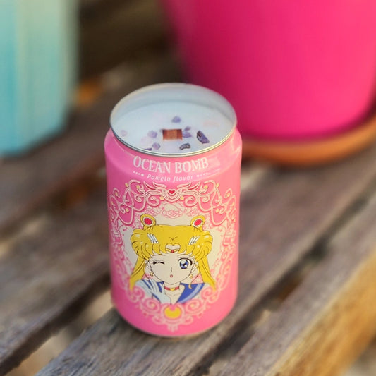 Eco-friendly candle made in a recycled Ocean Bomb soda can featuring a pink anime-themed design with a blonde character winking. The candle is topped with a wooden wick and purple crystals, sitting on a rustic wooden surface. Hand-poured with a coconut soy wax blend, this clean-burning, non-toxic candle adds a magical touch to any space and is perfect for fans of whimsical or manga-inspired decor.