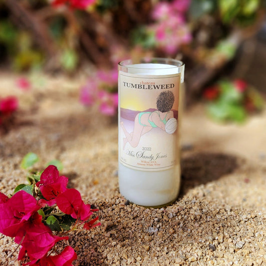 A hand-poured coconut soy wine bottle candle crafted from a recycled Chateau Tumbleweed Miss Sandy Jones bottle. The label features a playful beach volleyball illustration set against a warm desert sunset. The eco-friendly wooden wick candle has a smooth, polished glass rim, showcasing expert craftsmanship. Placed in a natural desert setting with vibrant pink bougainvillea flowers and sandy earth, this sustainable wine bottle candle is a perfect blend of Arizona charm and artisan luxury.