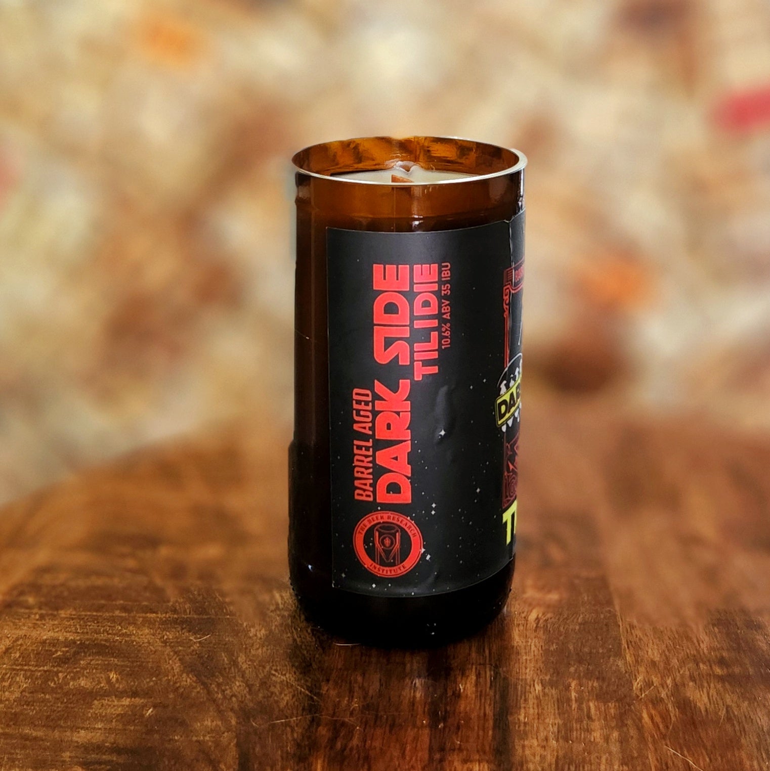 Recycled beer bottle candle showcasing the side label design, which includes bold red block lettering, “Dark Side Til I Die,” against a black background. A crackling wooden wick peeks through the white coconut soy wax.