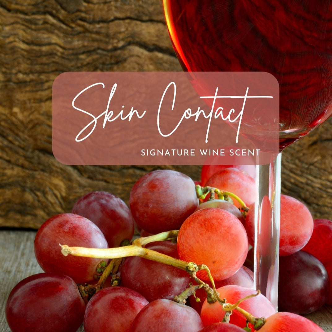 A close-up of plump, fresh red grapes resting on a wooden surface, paired with a partially visible wine glass filled with rich rose' wine. The background features a natural wood texture. Overlay text in an elegant script font reads "Skin Contact" with the subtitle "Signature Wine Scent," framed in a semi-transparent rectangle that complements the earthy and sophisticated aesthetic of the image.