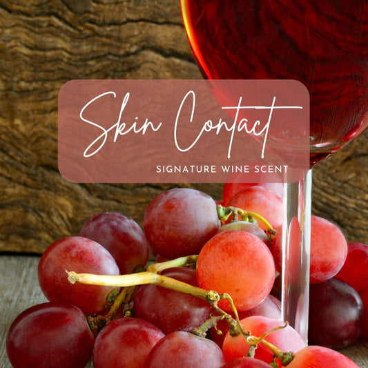 A close-up of plump, fresh red grapes resting on a wooden surface, paired with a partially visible wine glass filled with rich rose' wine. The background features a natural wood texture. Overlay text in an elegant script font reads "Skin Contact" with the subtitle "Signature Wine Scent," framed in a semi-transparent rectangle that complements the earthy and sophisticated aesthetic of the image.