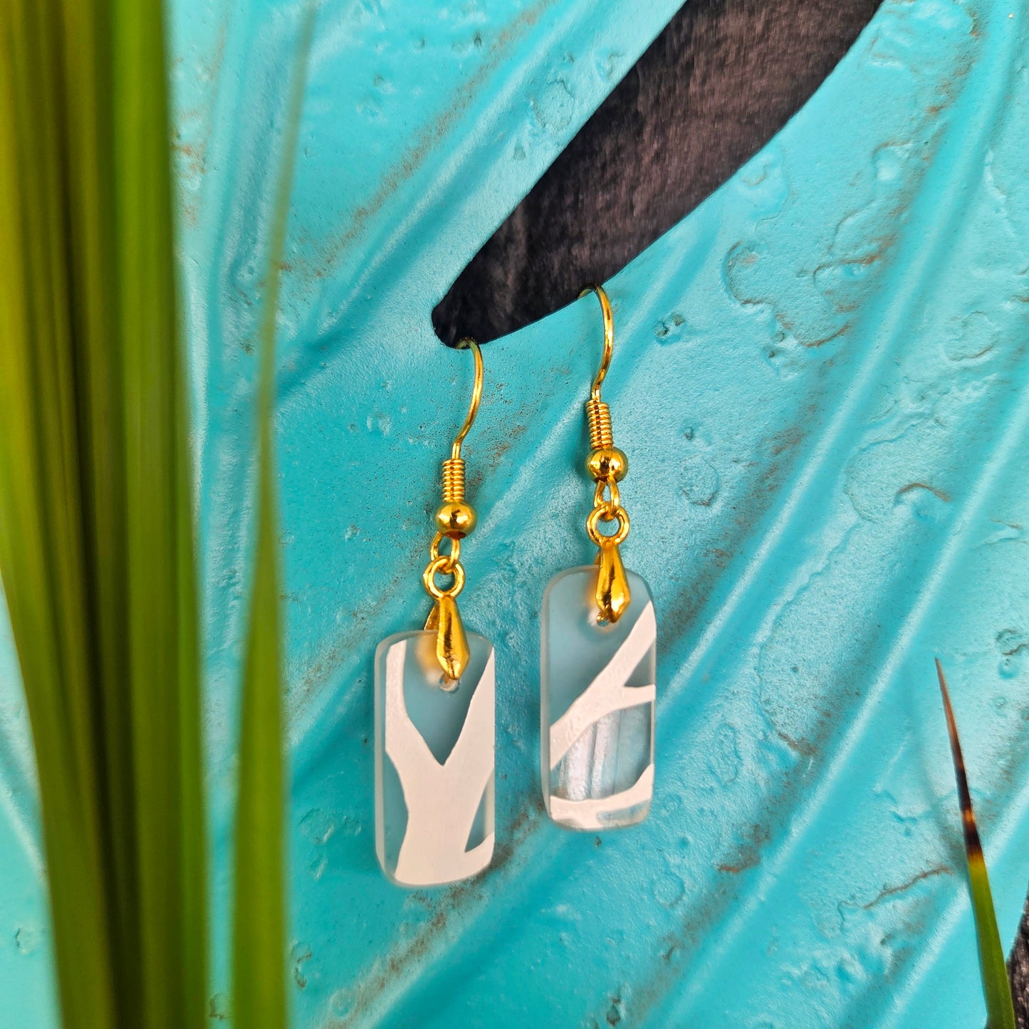 A pair of dangle earrings with rectangular translucent white and frosted clear glass, hanging against a bright turquoise textured background with blurred green leaves.