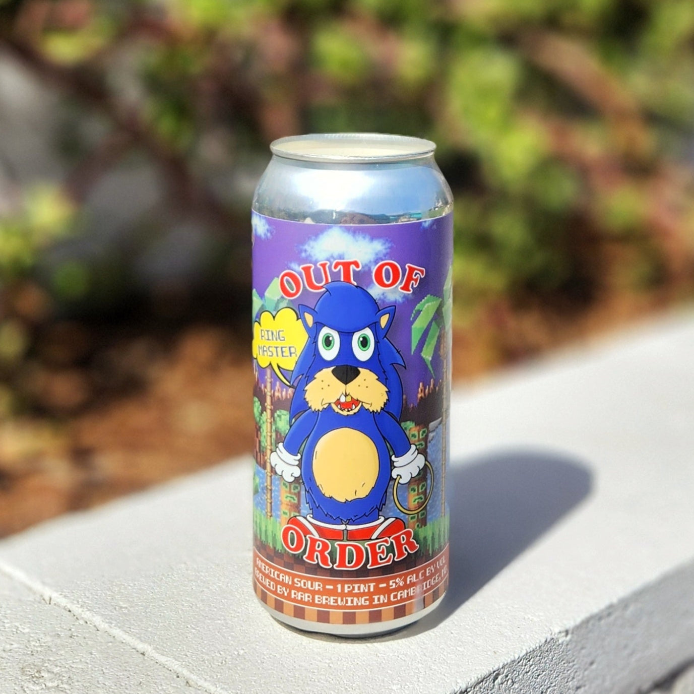 A hand-poured coconut soy wax candle housed in a recycled Out of Order beer can from RAR Brewing. The label features a humorous, cartoon-style parody of a well-known blue hedgehog, adding a playful and nostalgic touch. This eco-friendly wooden wick candle has a smooth, polished rim, showcasing expert craftsmanship. Placed on a sunlit white concrete ledge with blurred greenery in the background, this upcycled beer can candle is perfect for Sonic fans, craft beer lovers, and eco-conscious shoppers.