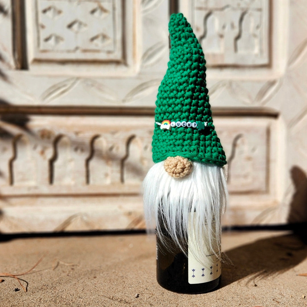 A crocheted gnome hat in green and white with a small beige pom-pom, placed on top of a dark bottle against a carved stone wall background.