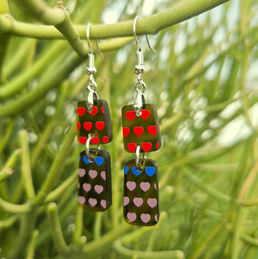A pair of dangling earrings with rectangular glass pendants featuring red and blue hearts on the top section and pink hearts on the bottom, hanging from silver fishhook wires against a blurred green foliage background.