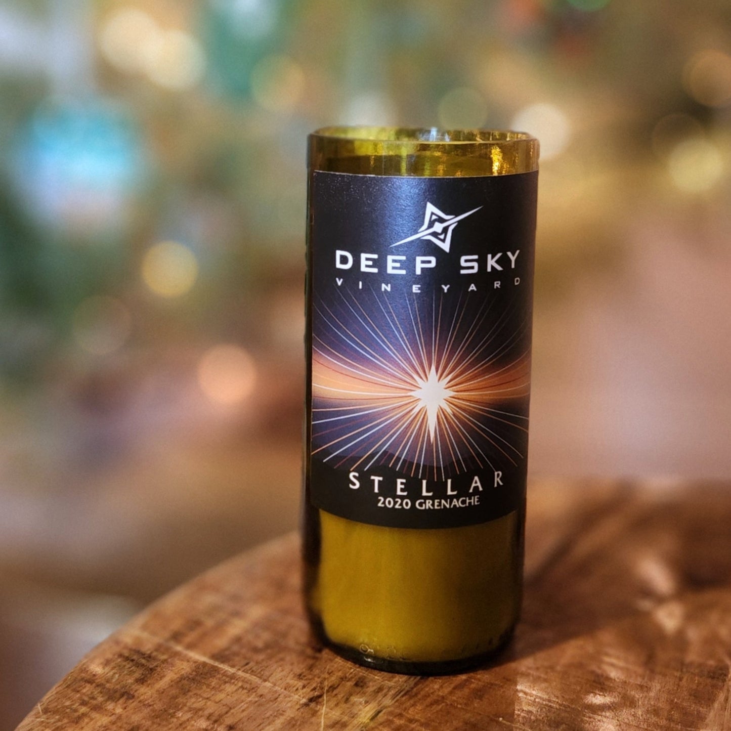 A hand-poured coconut soy wine bottle candle crafted from a recycled Deep Sky Vineyards bottle. The label showcases a striking celestial design with a radiant starburst, complementing the eco-friendly wooden wick candle. The polished glass rim reflects expert craftsmanship. Placed on a rustic wooden surface with a softly blurred holiday-lit background, this sustainable wine bottle candle is a perfect blend of luxury and sustainability for candle lovers and eco-conscious shoppers.