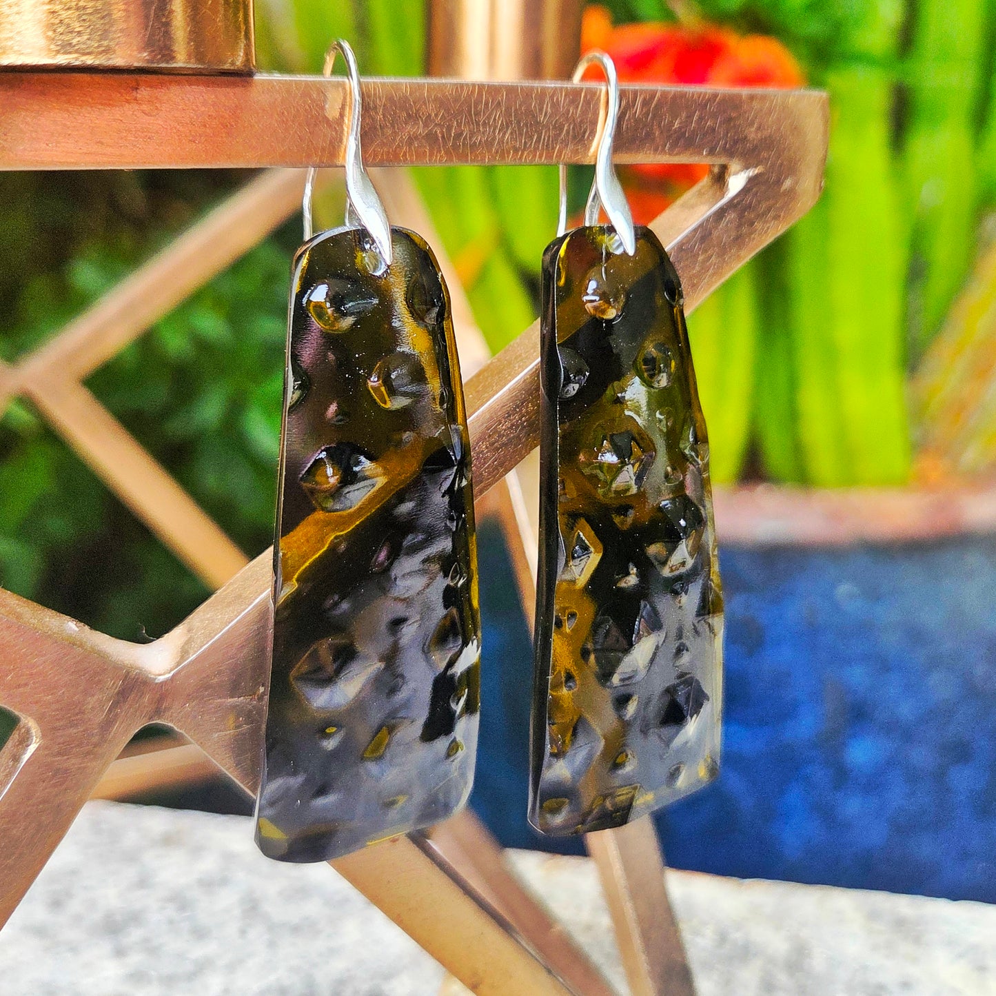 Pair of abstract glass earrings hanging on a metal stand with a blurred natural background