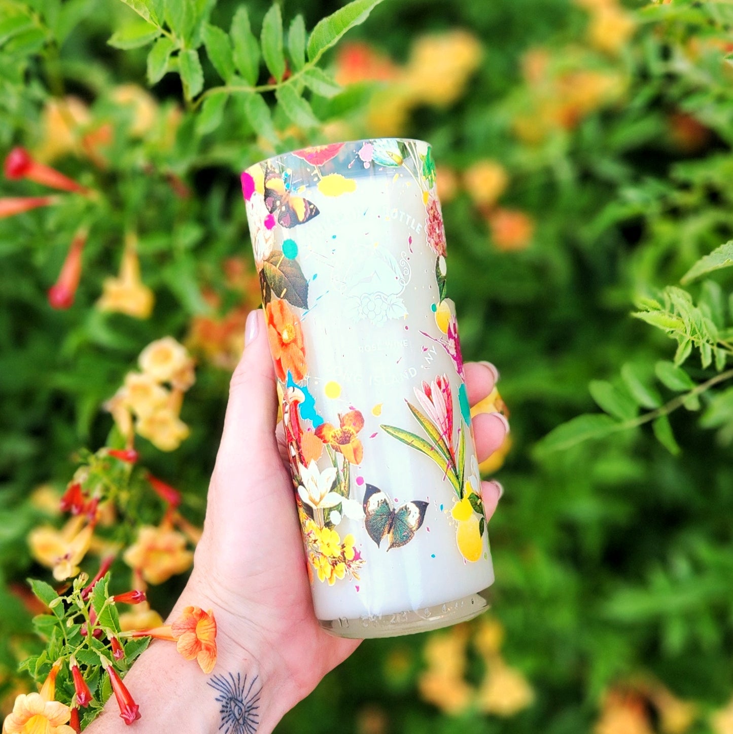 A vibrant eco-friendly wine bottle candle featuring the “Summer in a Bottle” floral and butterfly design, hand poured with coconut soy wax. Held outdoors in natural light, this upcycled bottle candle glows with colorful detailing and a clean, glossy rim.