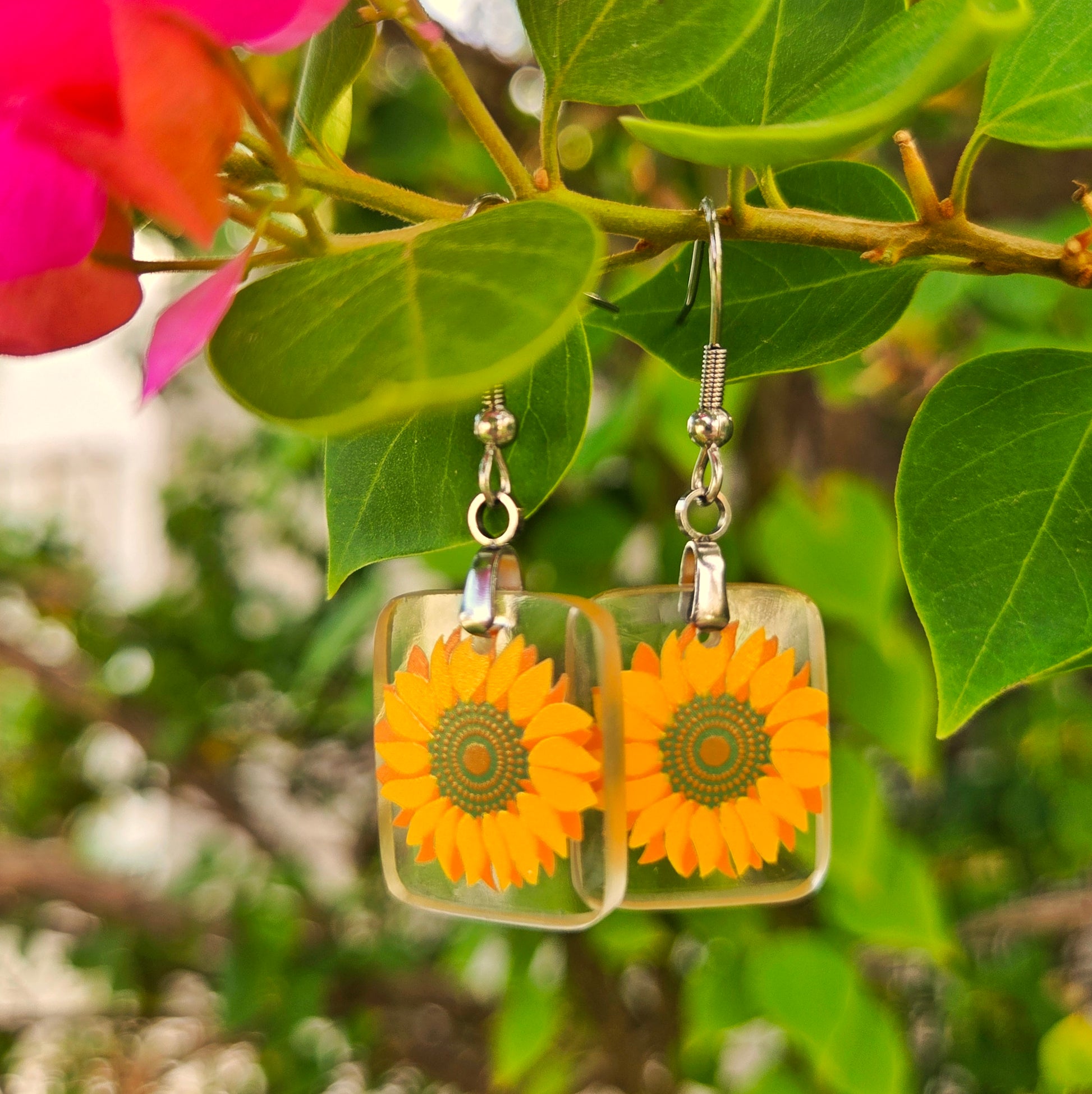 Square clear glass dangle earrings with printed orange-yellow sunflower designs, hanging from silver fishhook wires against green leaves and pink flowers.