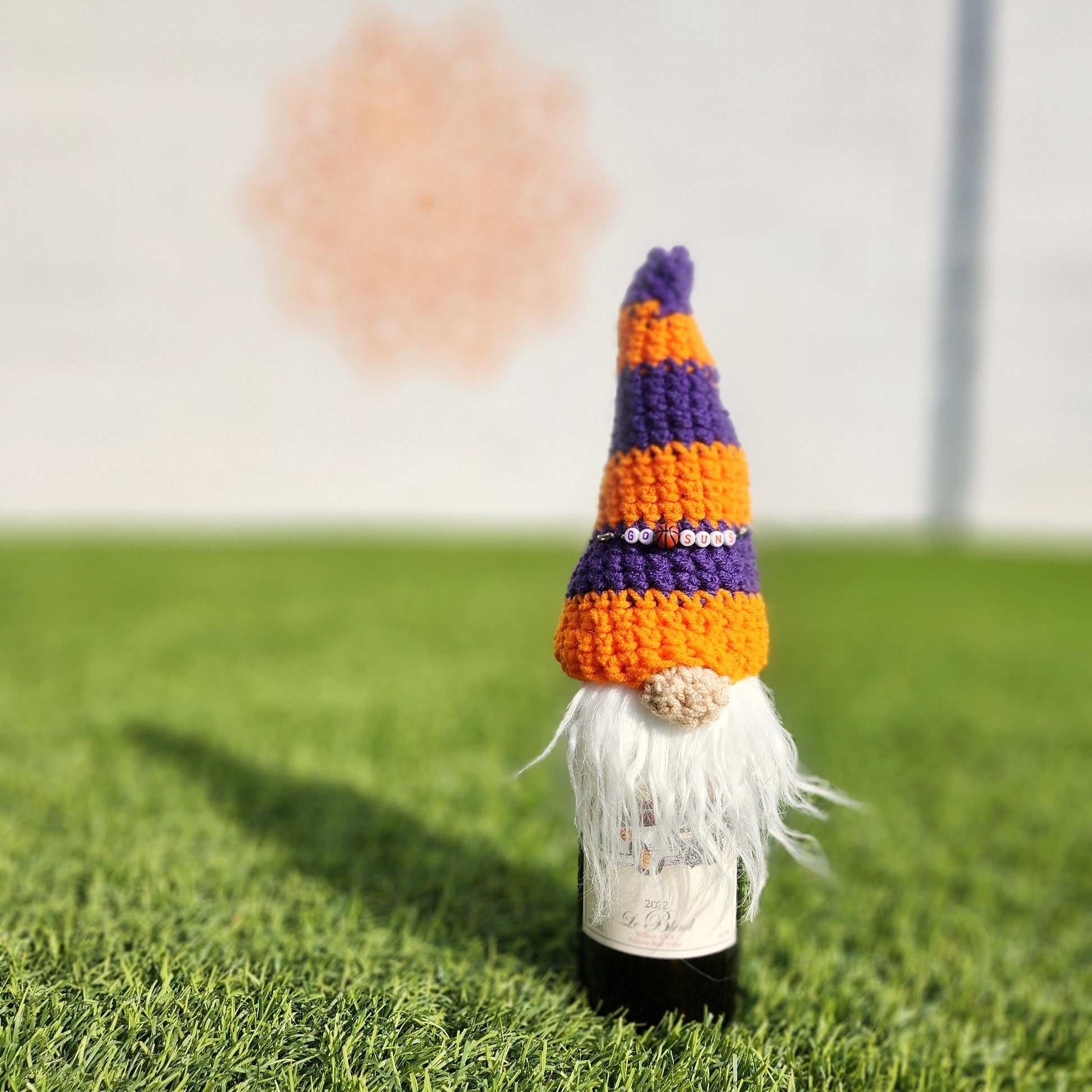 Purple and orange gnome bottle topper labeled “Go Suns” in beads, placed on a wine bottle on grass.

