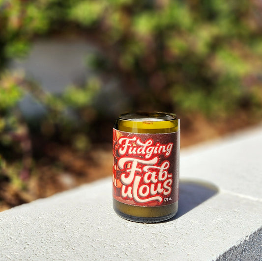 A hand-poured coconut soy wine bottle candle crafted from a recycled Superstition Meadery Fudging Fabulous bottle. The label features a bold, vintage-style design with rich red and white lettering, reflecting the indulgent chocolate mead theme. This eco-friendly wooden wick candle has a polished glass rim, showcasing expert craftsmanship. Placed outdoors on a sunlit concrete ledge with a softly blurred green backdrop.