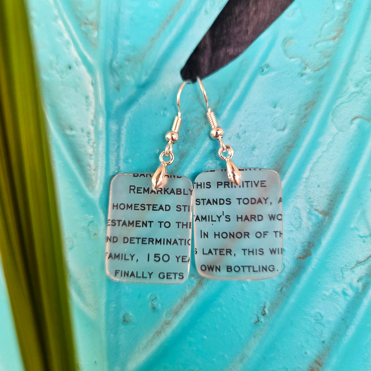 A pair of handmade dangle earrings with rectangular frosted glass pendants printed with black text, hanging from silver-toned fishhook wires against a textured turquoise background.