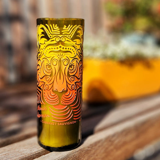 A recycled wine bottle candle with a smooth glass rim, featuring an intricate etched design of a tiger's face in vibrant orange and yellow gradients. The label reads "Manos Artisan Cabernet Sauvignon." The candle is set on a wooden bench with a colorful blurred background of planters and flowers, creating a bold and artistic outdoor display.