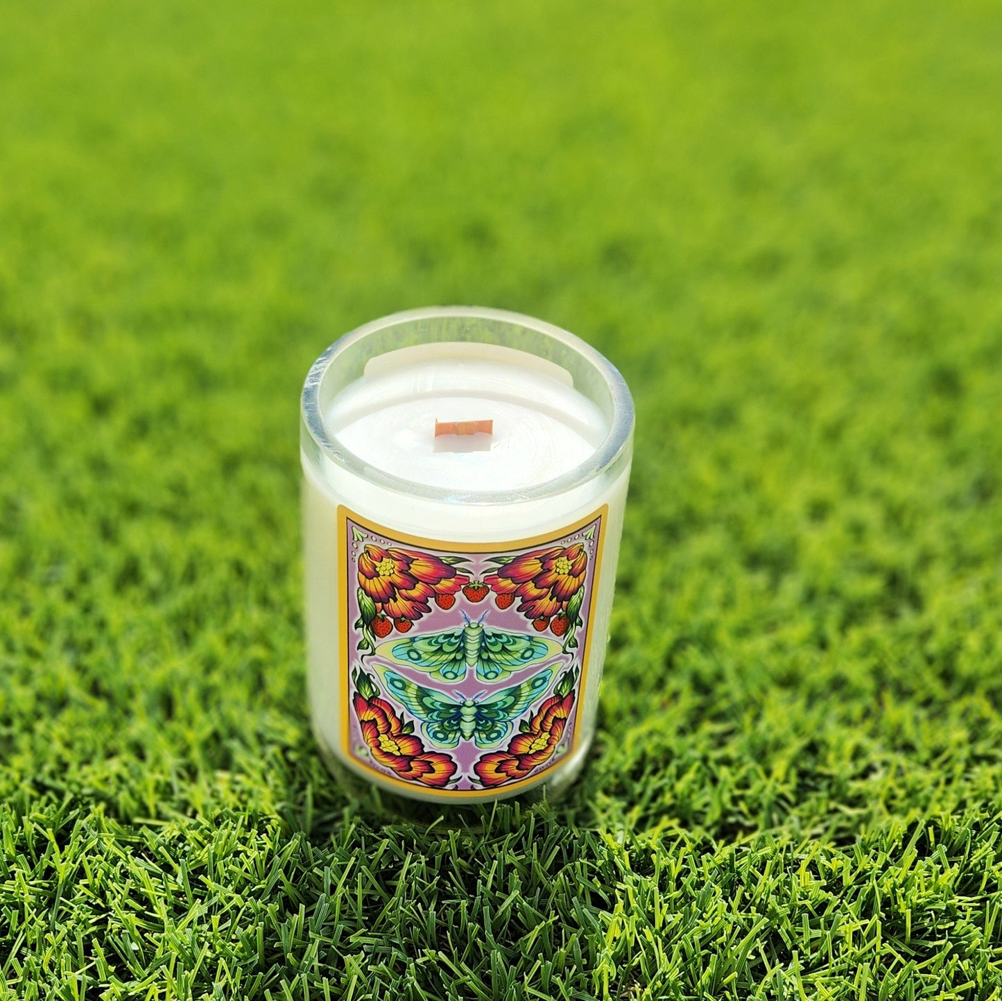 Eco-friendly coconut soy candle in a recycled wine bottle with a bright, psychedelic-style label featuring a green and orange design. The wooden wick is centered in the creamy wax, framed by a shiny, smooth glass rim. Photographed on vibrant green grass.