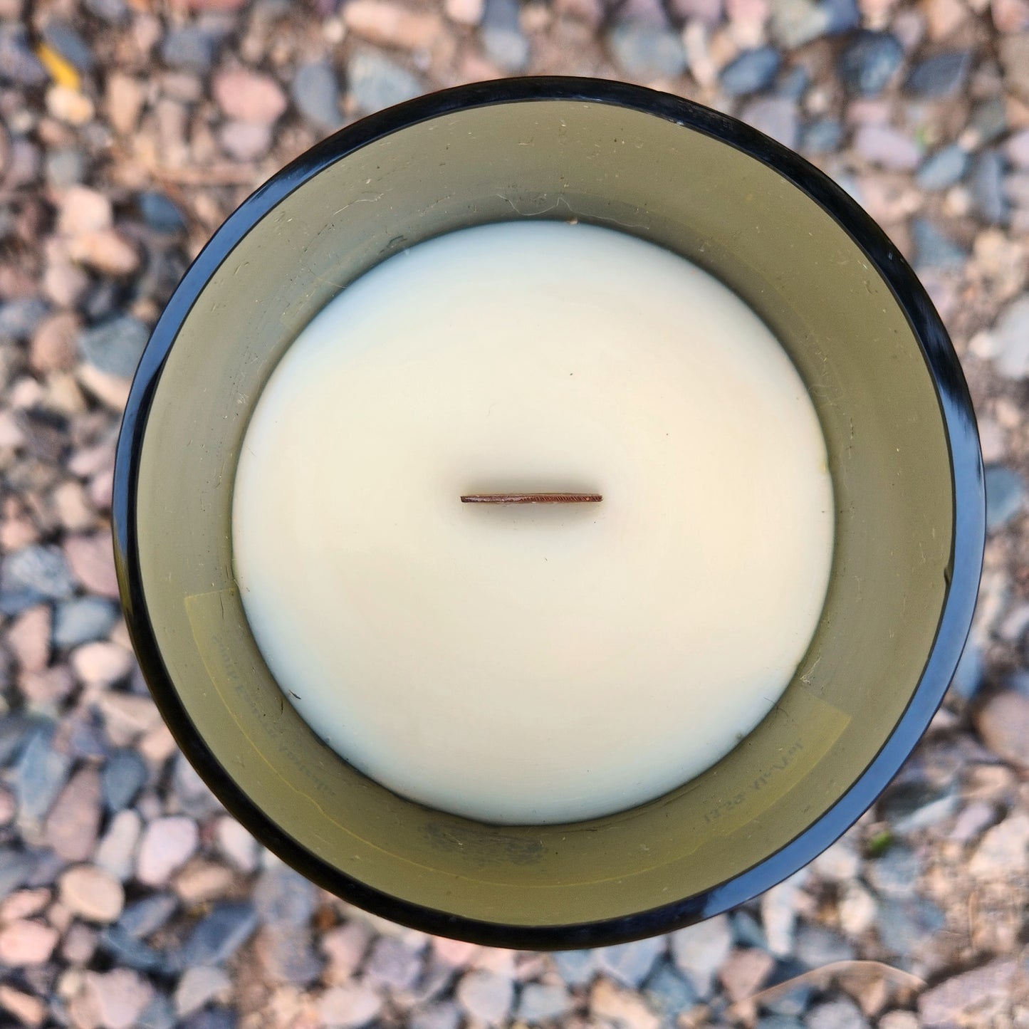 Top-down view of an eco-friendly coconut soy candle housed in a recycled wine bottle with a smooth, polished glass rim. The creamy white wax is topped with a wooden wick, centered precisely for an even burn. The translucent green glass vessel subtly reflects light, and the candle rests on a bed of gravel, enhancing its earthy, sustainable aesthetic.