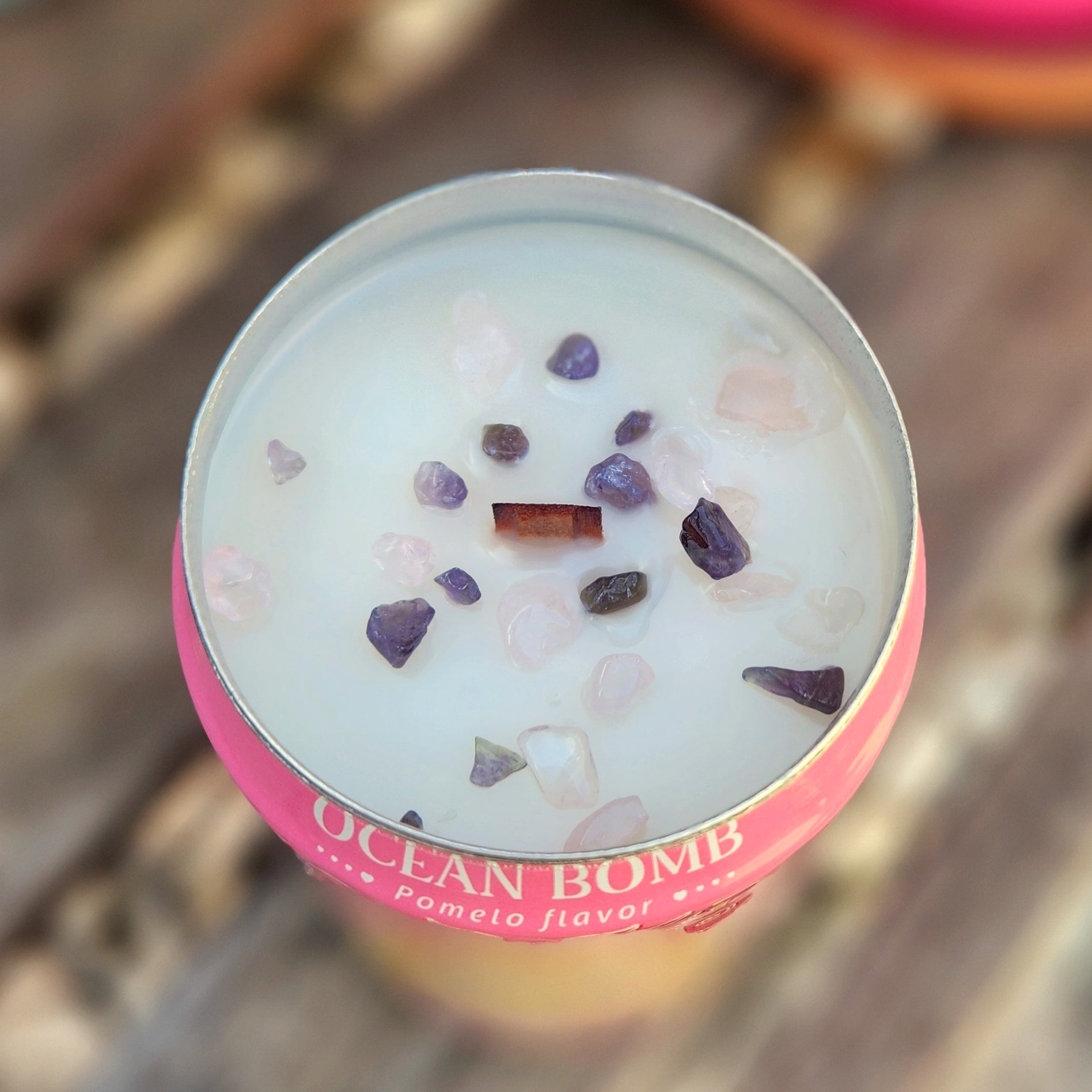 A top-down view of a pink Ocean Bomb can candle with a wooden wick and scattered rose quartz and amethyst crystal pieces embedded in creamy coconut soy wax.