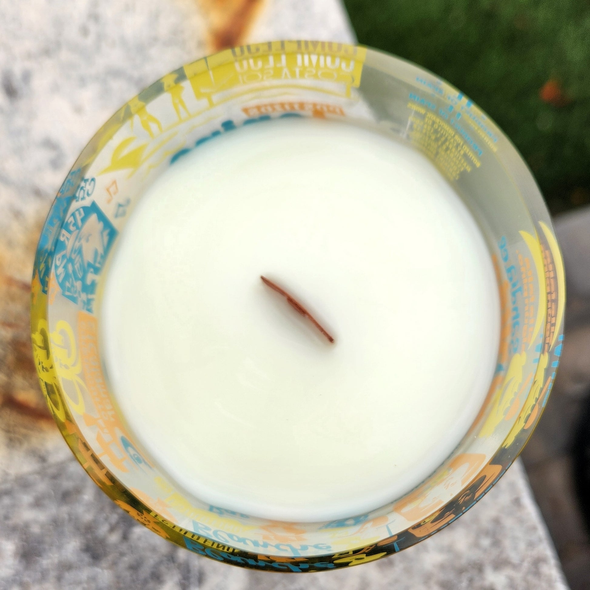 Top-down view of an unlit eco-friendly wine bottle candle showing creamy coconut soy wax and a centered wooden wick; the surrounding glass is covered in bold, vintage-style illustrations.