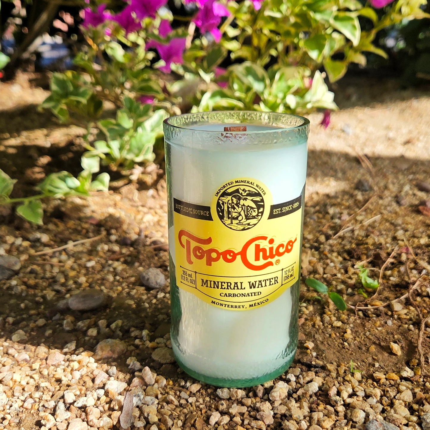 Topo Chico mineral water bottle repurposed as a candle on a gravel surface with plants in the background