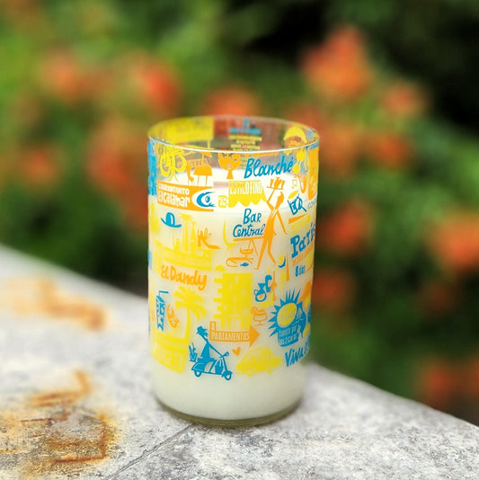 Another side of the recycled candle vessel reveals bold Spanish travel-themed artwork and colorful graphics wrapping around the clear glass; lush green garden background.