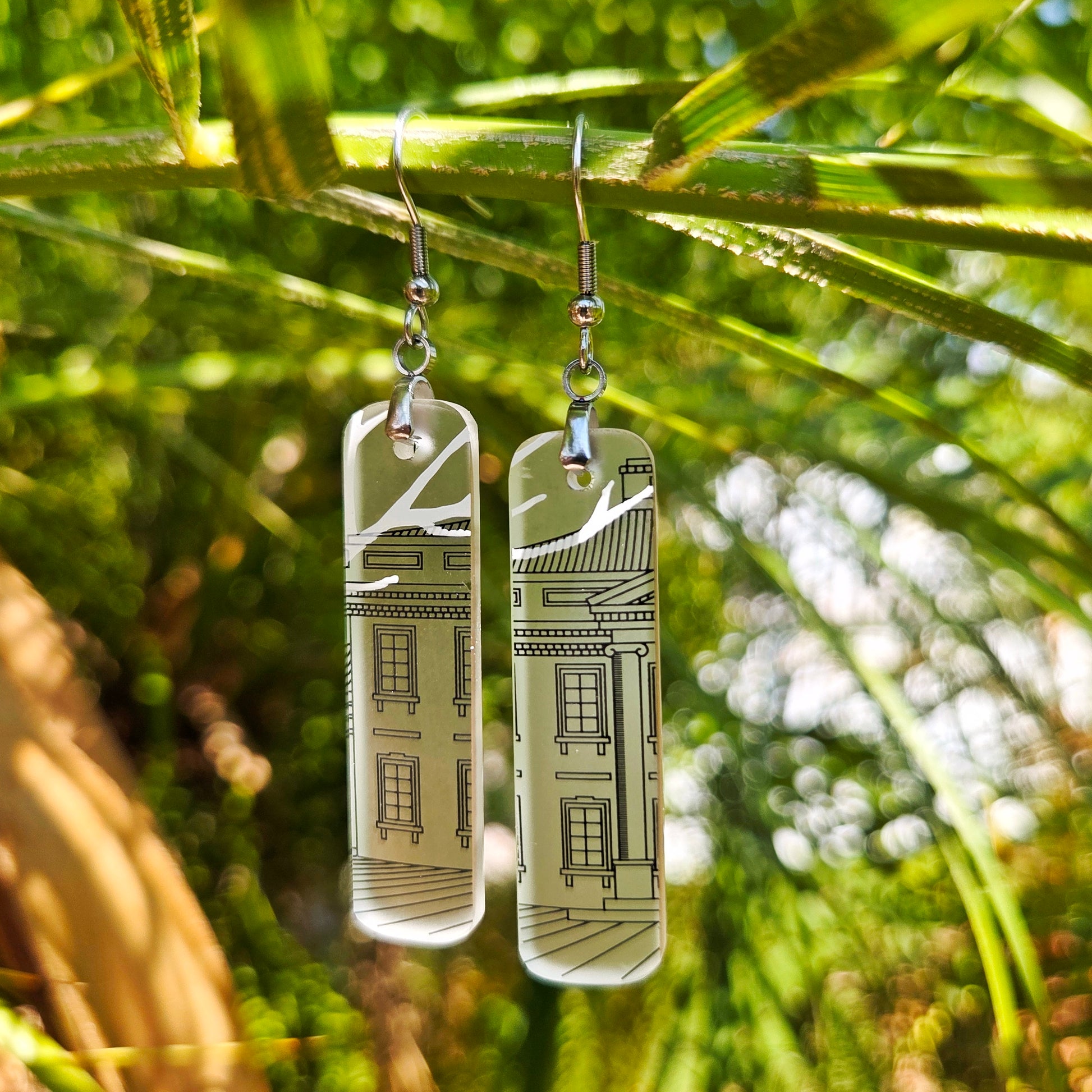 A pair of rectangular clear glass drop earrings with a black architectural building design, hanging from silver fishhook wires. The earrings are photographed outdoors hanging from a green palm branch with a blurred natural background.