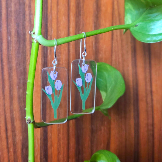 Transparent earrings with floral design hanging on a green branch against a wooden background