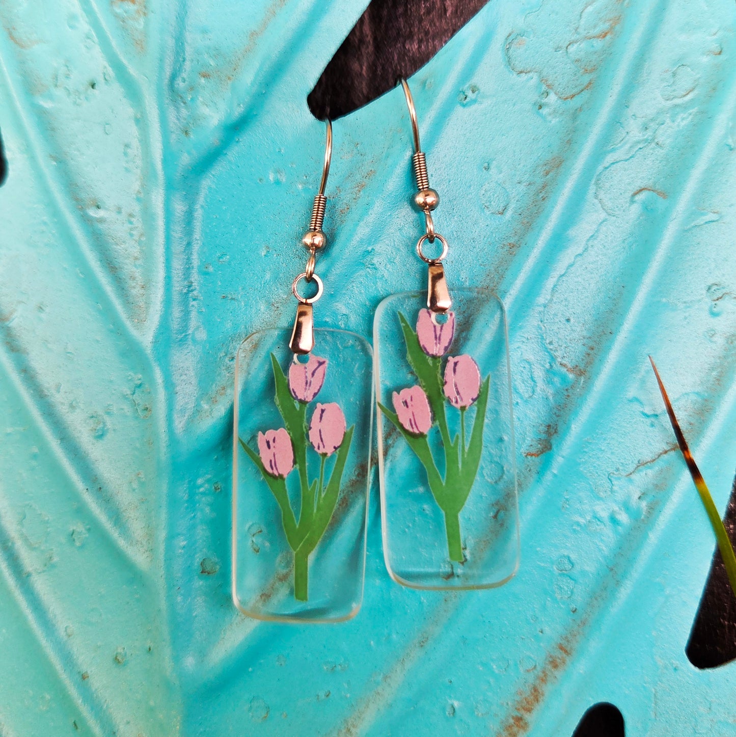 A pair of rectangular clear glass drop earrings featuring purple tulips with green stems, suspended from gold-toned wire hooks, displayed against a textured turquoise leaf background.