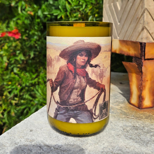 Wine bottle candle featuring art of a cowgirl with lasso.  Candle is sitting on a stone surface with a rusted planter and greenery in the background.
