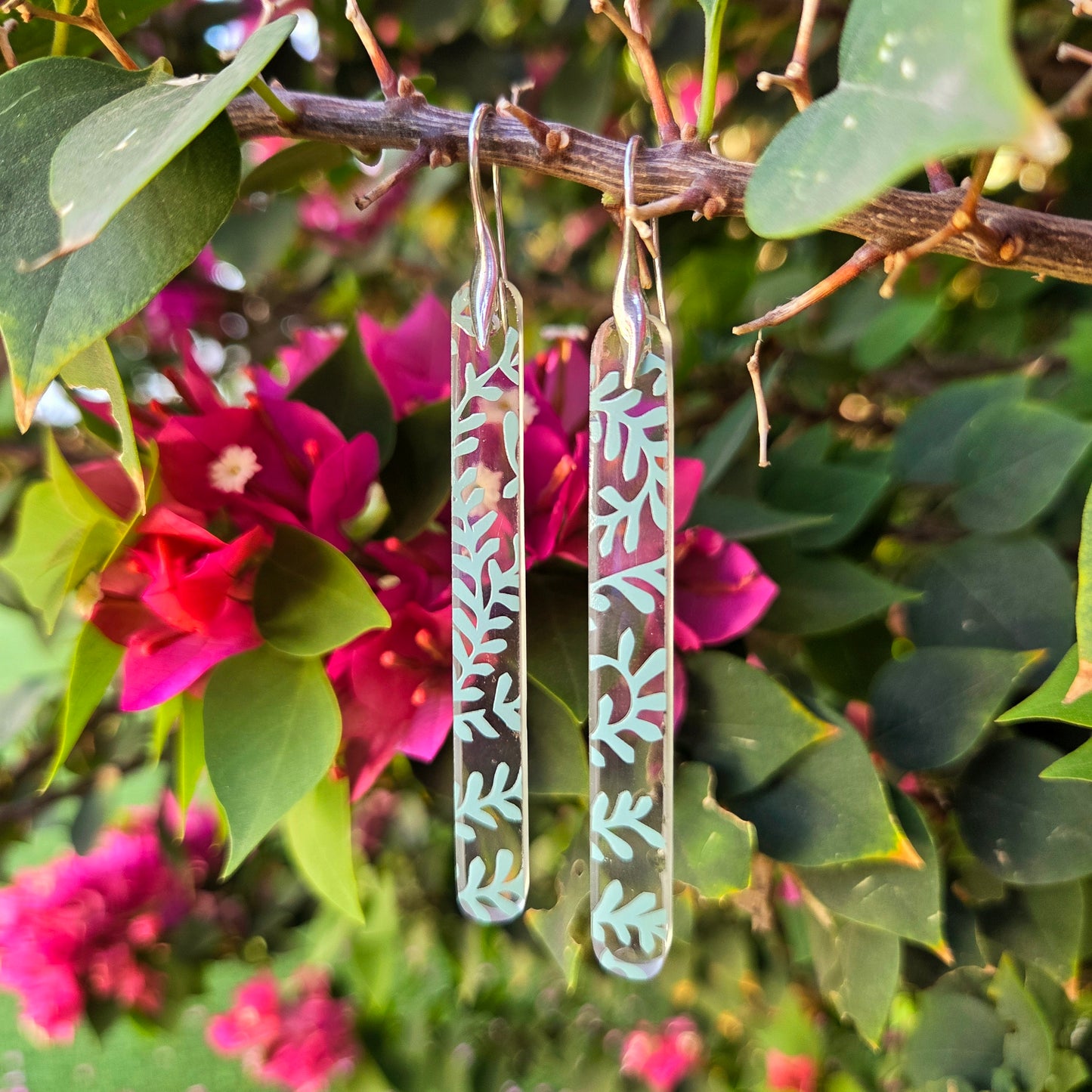 Clear glass vine-patterned earrings made from reclaimed gin bottles, hanging on a branch with pink flowers and green leaves.