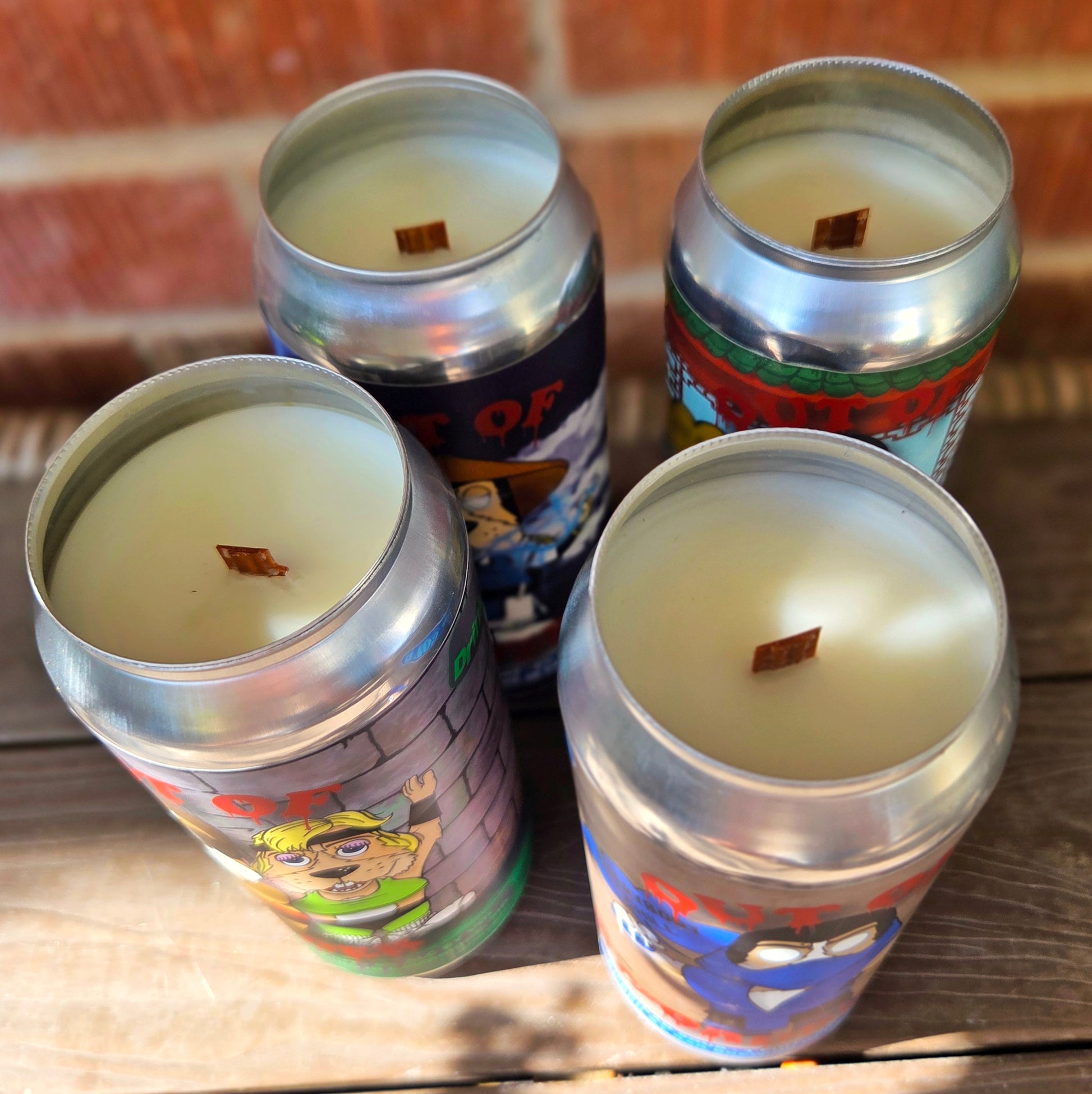 Overhead view of four handcrafted beer can candles, each filled with smooth coconut soy wax and a centered wooden wick. The cans feature colorful Mortal Kombat-inspired cartoon dog characters, making these eco-friendly, upcycled candles both collectible and functional.