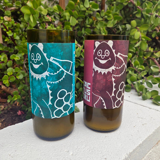 Two wine bottle candles with colorful labels featuring cartoon-like designs on a white surface with greenery in the background.