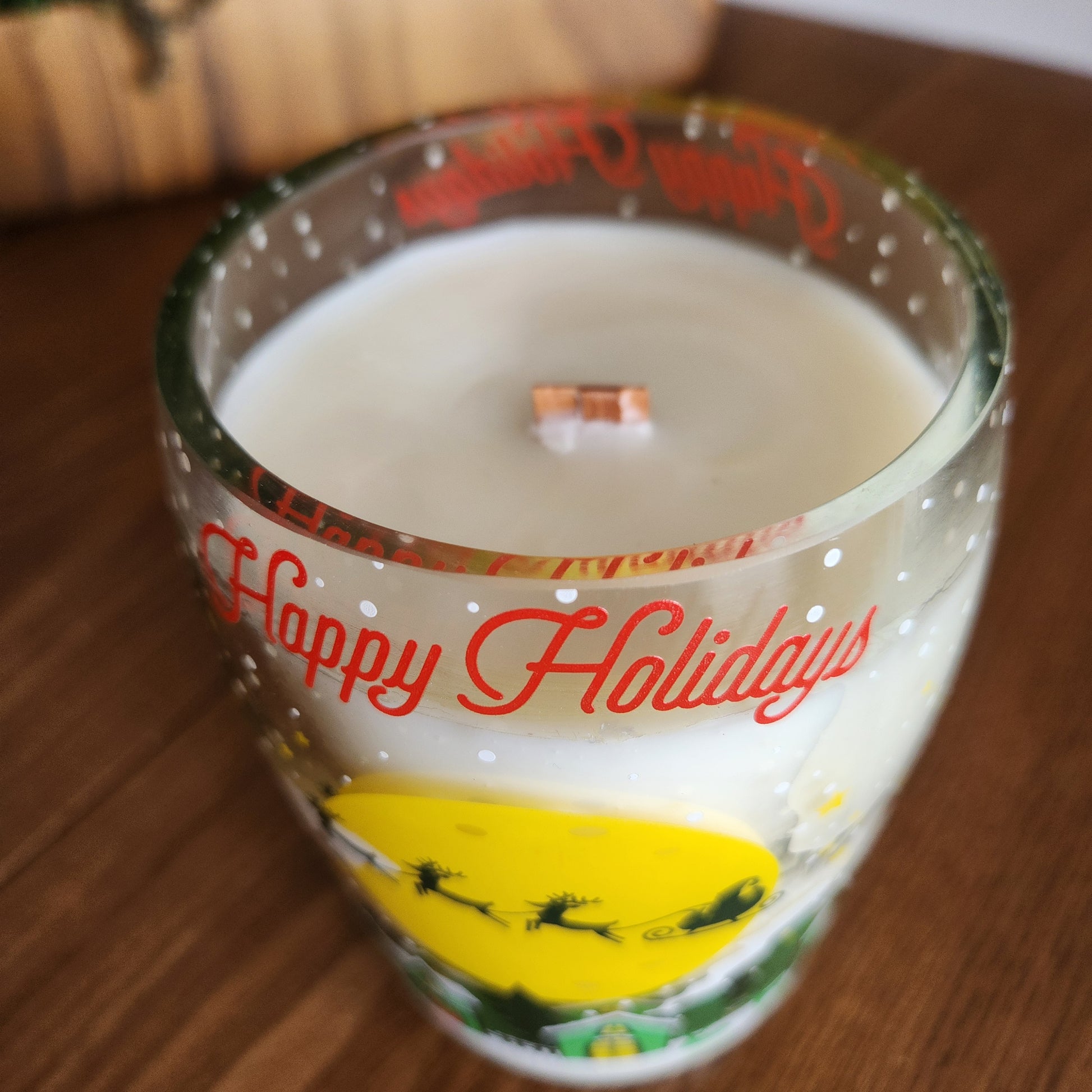 Angled view of a festive wine bottle candle featuring “Happy Holidays” text and Santa's sleigh artwork, hand-poured with coconut soy wax and a wooden wick in a recycled glass vessel.