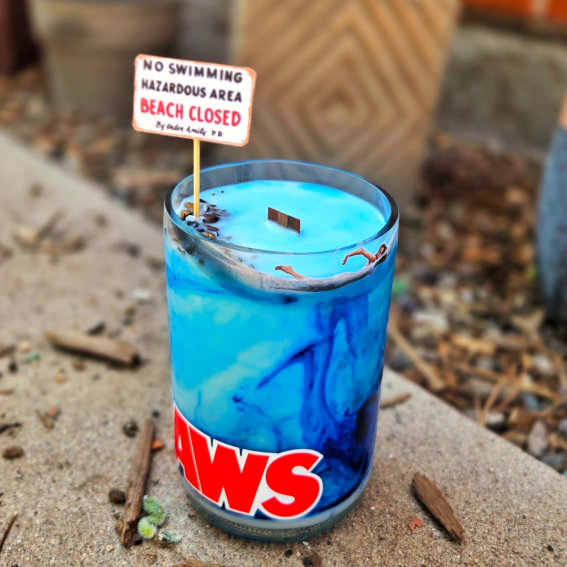 Angled shot of a recycled wine bottle candle featuring a Jaws label, swirling blue wax, tiger's eye crystals, a wooden wick, and a mini beach warning sign—evoking coastal danger in a fun, oceanic design.