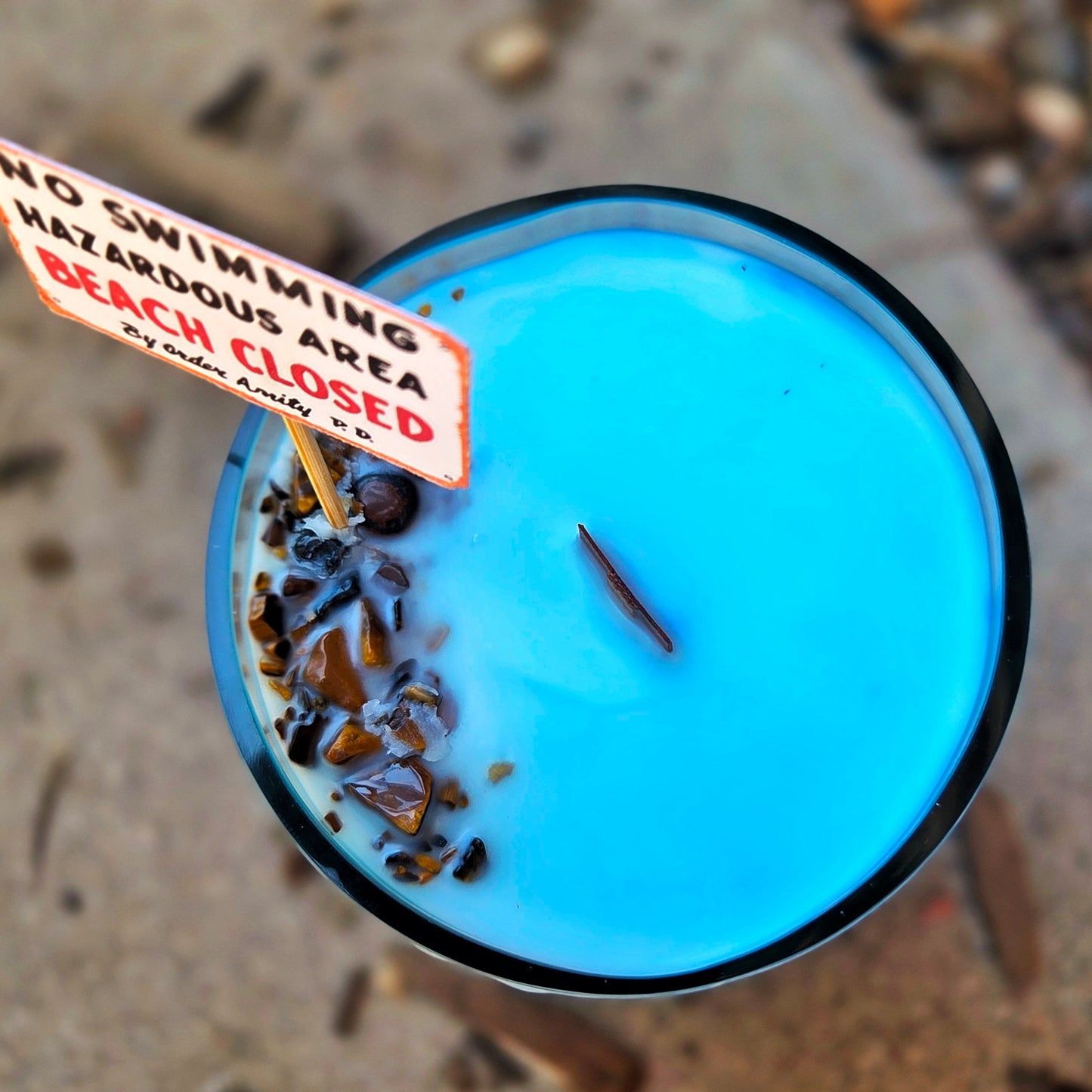 Top view of a Jaws-inspired wine bottle candle with bright blue coconut soy wax, a centered wooden wick, and tumbled tiger's eye crystals embedded on the surface beside a mini "Beach Closed" warning sign.