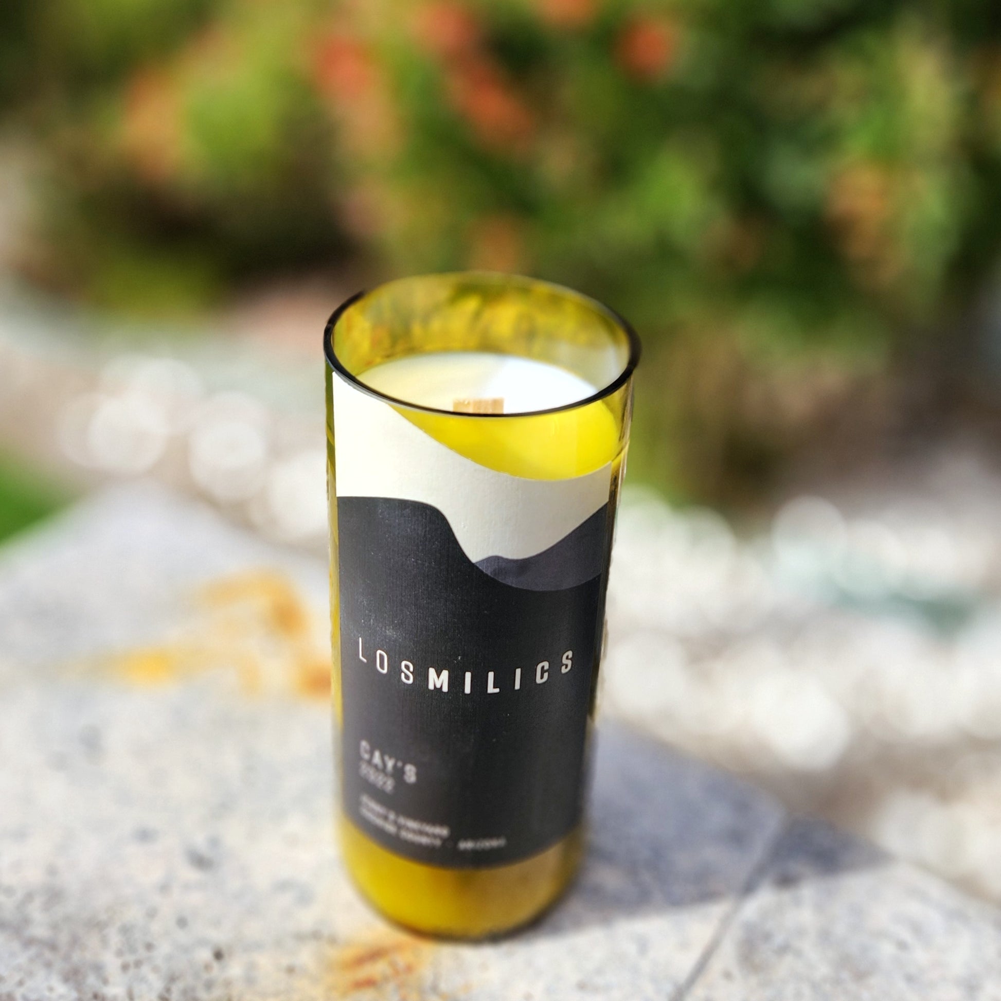 Eco-friendly candle hand poured into a recycled Los Milics wine bottle with bold black and white abstract label. Features smooth cut rim and wooden wick, set outdoors in natural light.