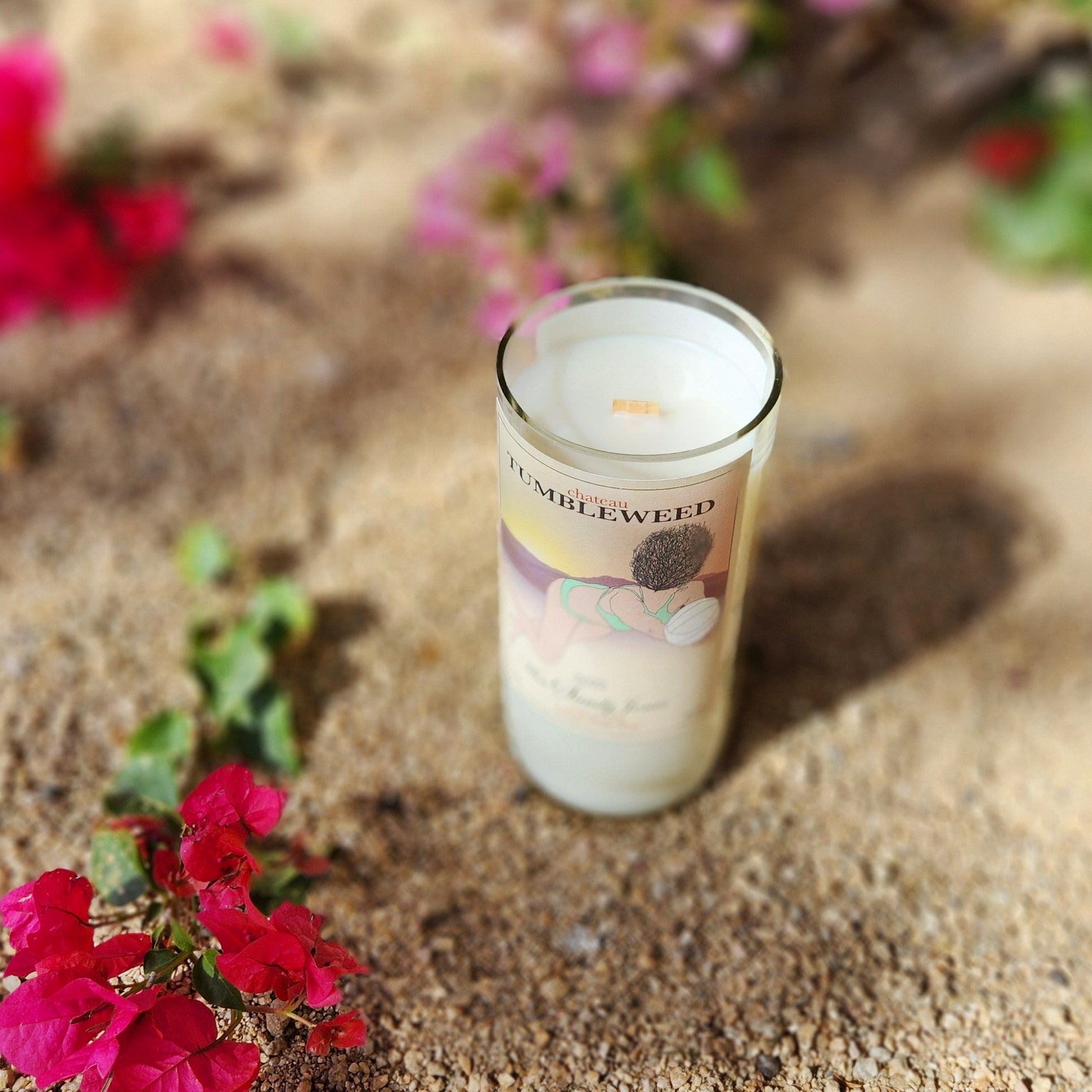 Chateau Tumbleweed wine bottle candle in the Sandy Jones design, sitting on sunlit desert sand with pink bougainvillea flowers in the background.