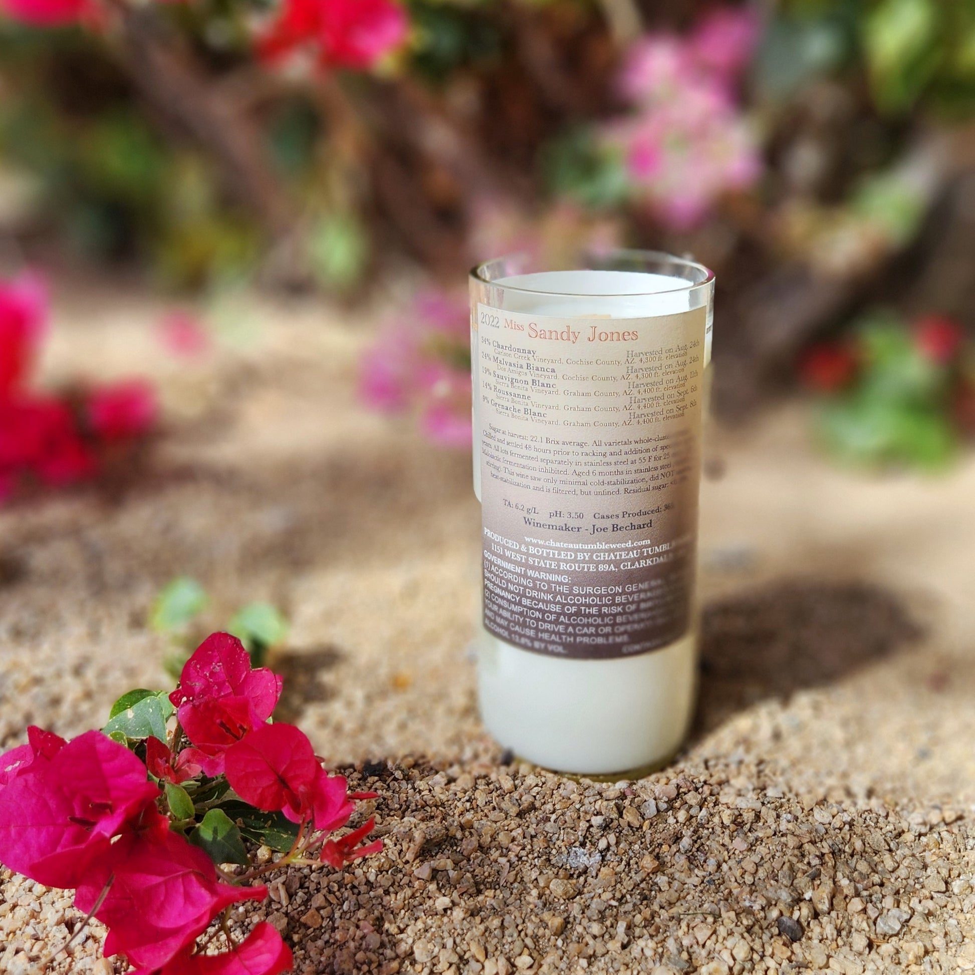 Recycled Sandy Jones wine bottle candle showing detailed vineyard back label, with creamy wax and floral garden backdrop for a natural, handmade vibe.