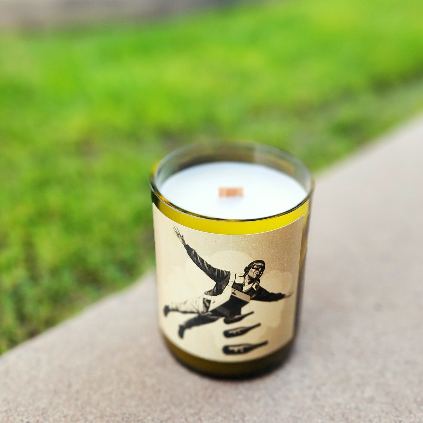 A handmade wine bottle candle with a striking label featuring a parachuting figure and falling bombs, placed on a stone ledge against a vivid green lawn. The clean-burning coconut soy wax and wooden wick sit smoothly inside the upcycled bottle vessel.