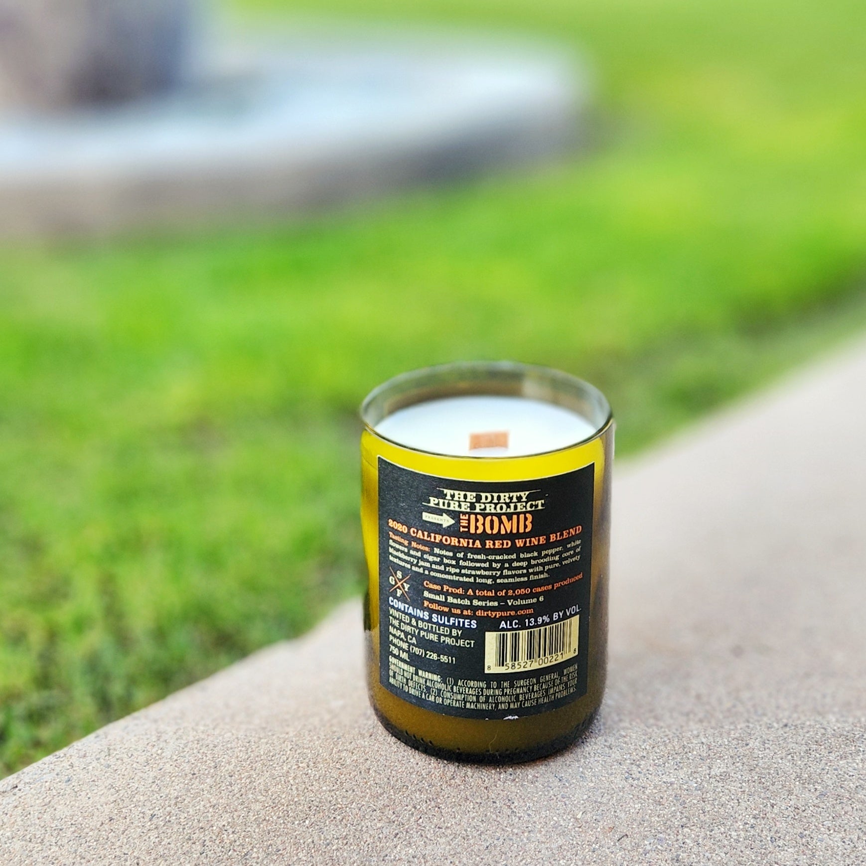 A recycled wine bottle candle displayed outdoors on a curved stone ledge with vibrant grass in the background. The back label reads “The Dirty Pure Project: The Bomb,” identifying the wine as a 2020 California red blend. The white coconut soy wax and wooden wick are clearly visible.