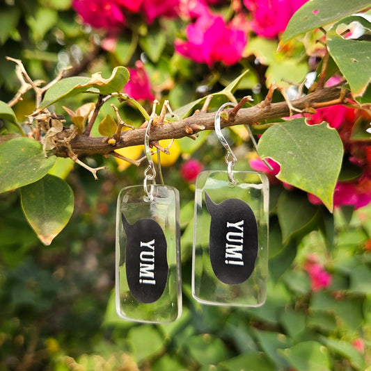 Clear glass earrings with black speech bubble saying yum. hanging on a branch with pink flowers in the background