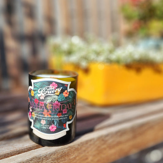 A recycled glass candle crafted from a dark green beer bottle, featuring a vibrant label with tropical elements, including palm leaves and colorful hibiscus flowers. The label reads "Aloha Friday" with branding from "The Bruery" and details about the original beverage. The candle sits on a wooden surface outdoors, with a yellow planter holding greenery in the blurred background, creating a warm and inviting atmosphere.