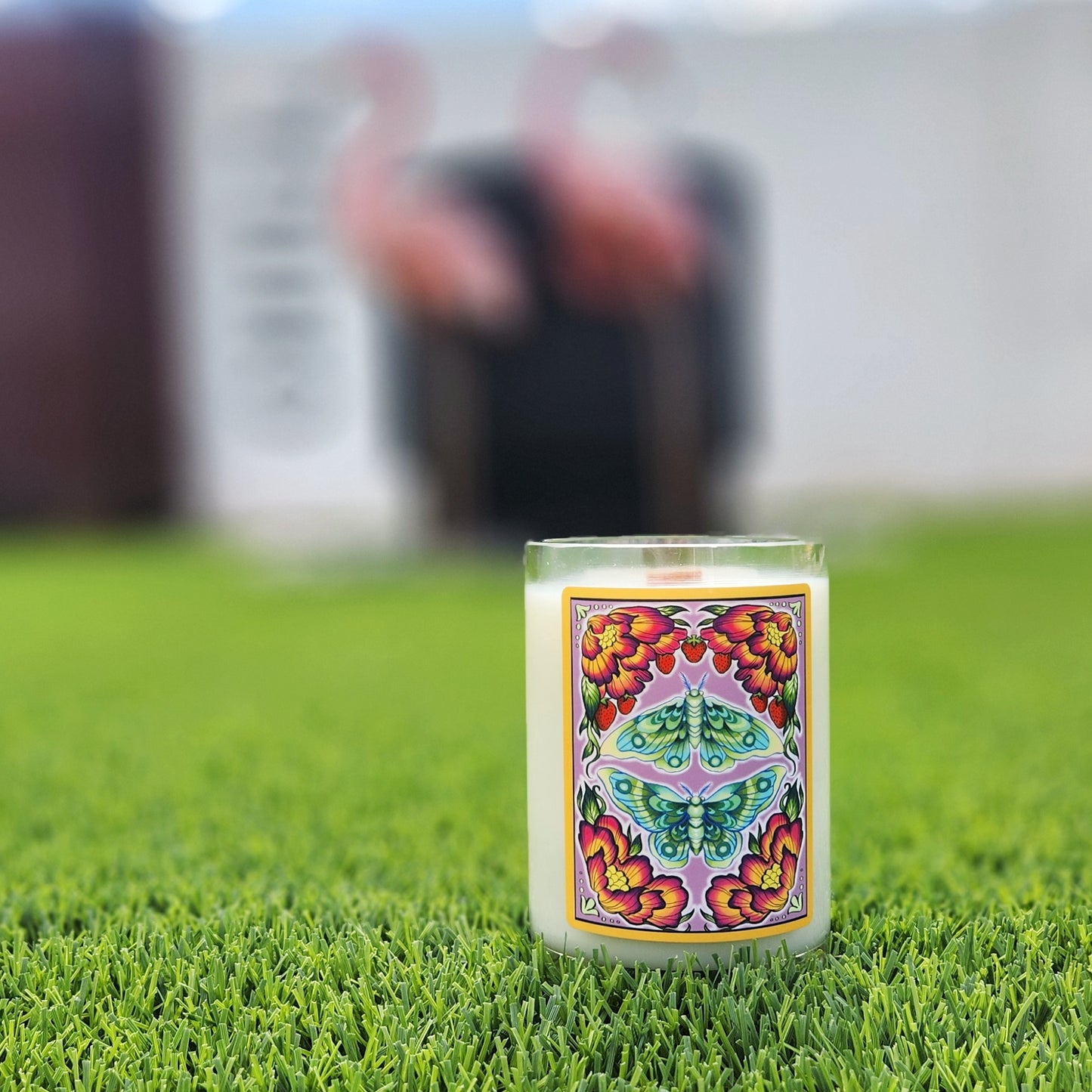 A handcrafted candle housed in a recycled glass mead bottle, featuring a vibrant label with a symmetrical design of a green luna moth surrounded by blooming red and yellow flowers and delicate strawberries. The candle sits on lush green grass, with a blurred background of pink flamingo yard decorations, creating a whimsical and artistic outdoor setting. The smooth glass rim and creamy white wax enhance its artisanal and eco-friendly appeal.