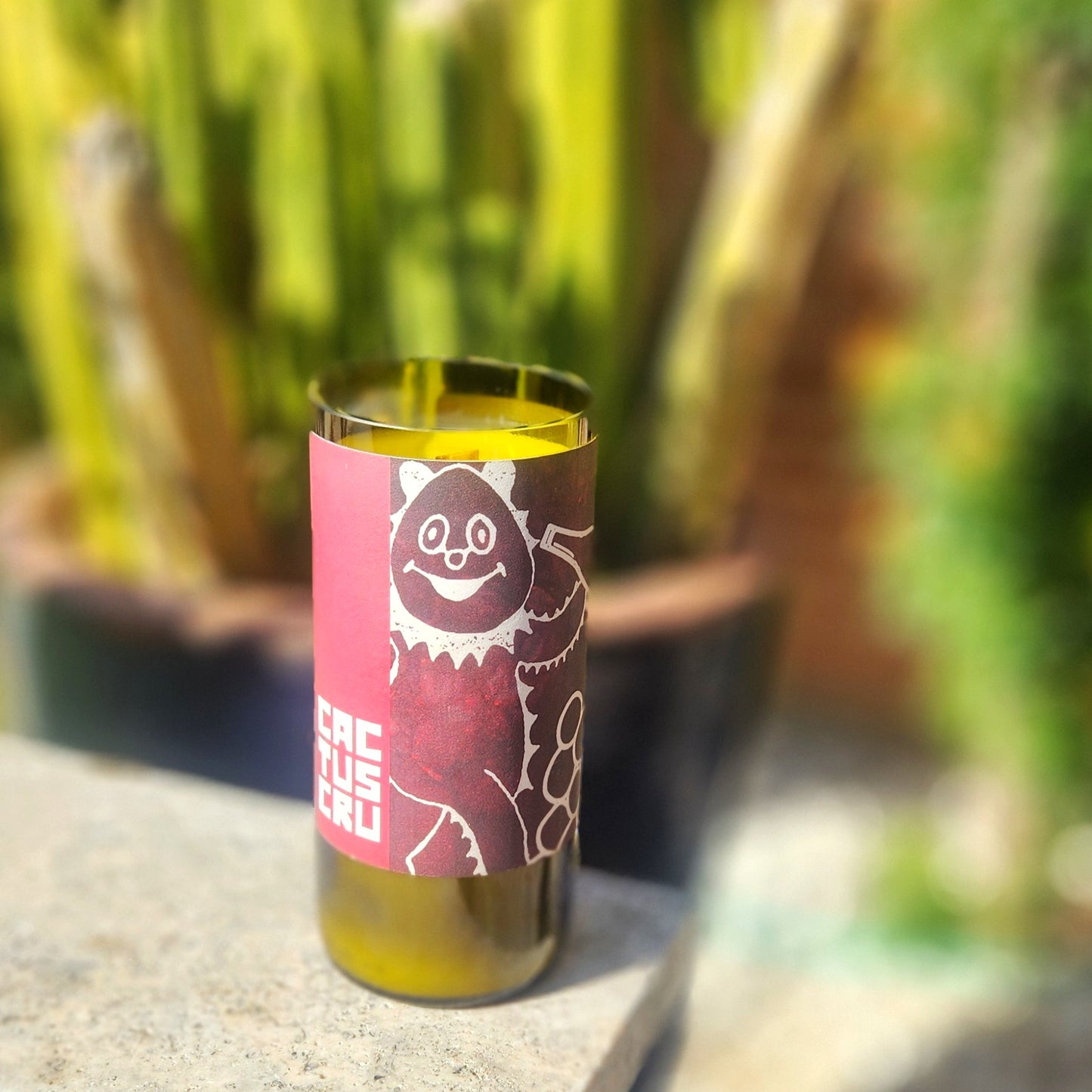 A recycled glass candle made from a green wine bottle, featuring a playful red and white label with a whimsical design of a smiling cactus figure. The label reads "Cactus Cru" in bold text. The candle is outdoors, resting on a light-colored stone surface, with a blurred background of potted cacti and greenery, evoking a cheerful, desert-inspired atmosphere.