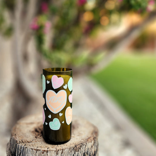 A hand-poured candle made from coconut-soy wax in a recycled green wine bottle with a shiny, smooth rim. The candle features a playful design of pastel-colored candy hearts with phrases like "XOXO," "Be Mine," and "I Adore You," evoking a Valentine’s Day theme. It rests on a rustic tree stump outdoors, with a blurred background of vibrant greenery and blooming pink flowers, creating a romantic and festive atmosphere. Perfect for eco-friendly holiday gifting or decor.

