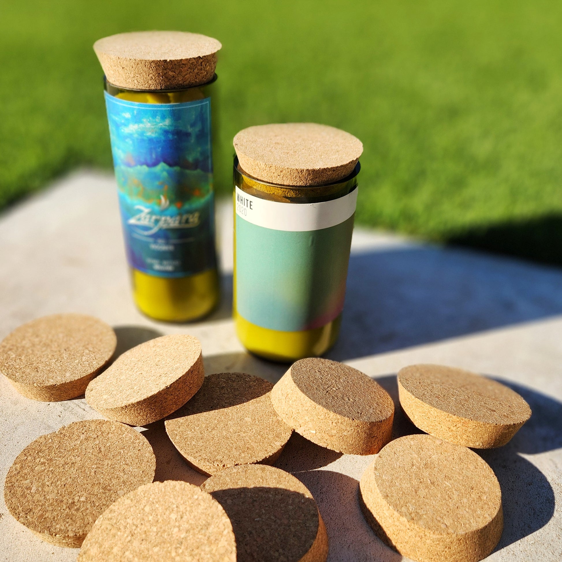 Two recycled wine bottle candles topped with round cork lids are displayed on a concrete surface, surrounded by additional loose cork lids. One candle features a vibrant blue and green label, while the other has a minimalist design with a soft gradient. A lush green grass background adds a natural, eco-friendly vibe to the composition.