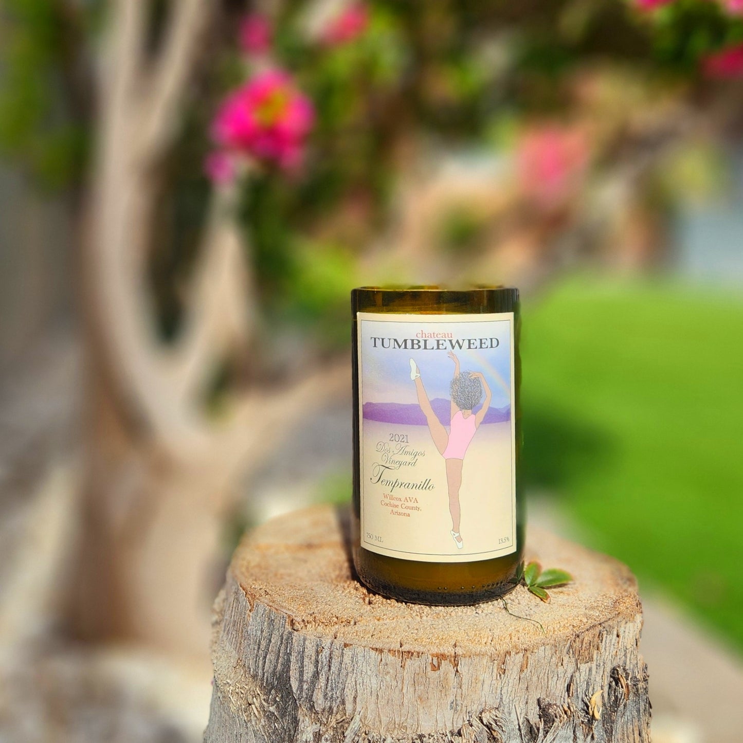 Elegant and eco-friendly, this hand-poured candle is crafted in a recycled Chateau Tumbleweed wine bottle. Set on a rustic tree stump with a serene outdoor backdrop, it combines sustainability with natural charm.