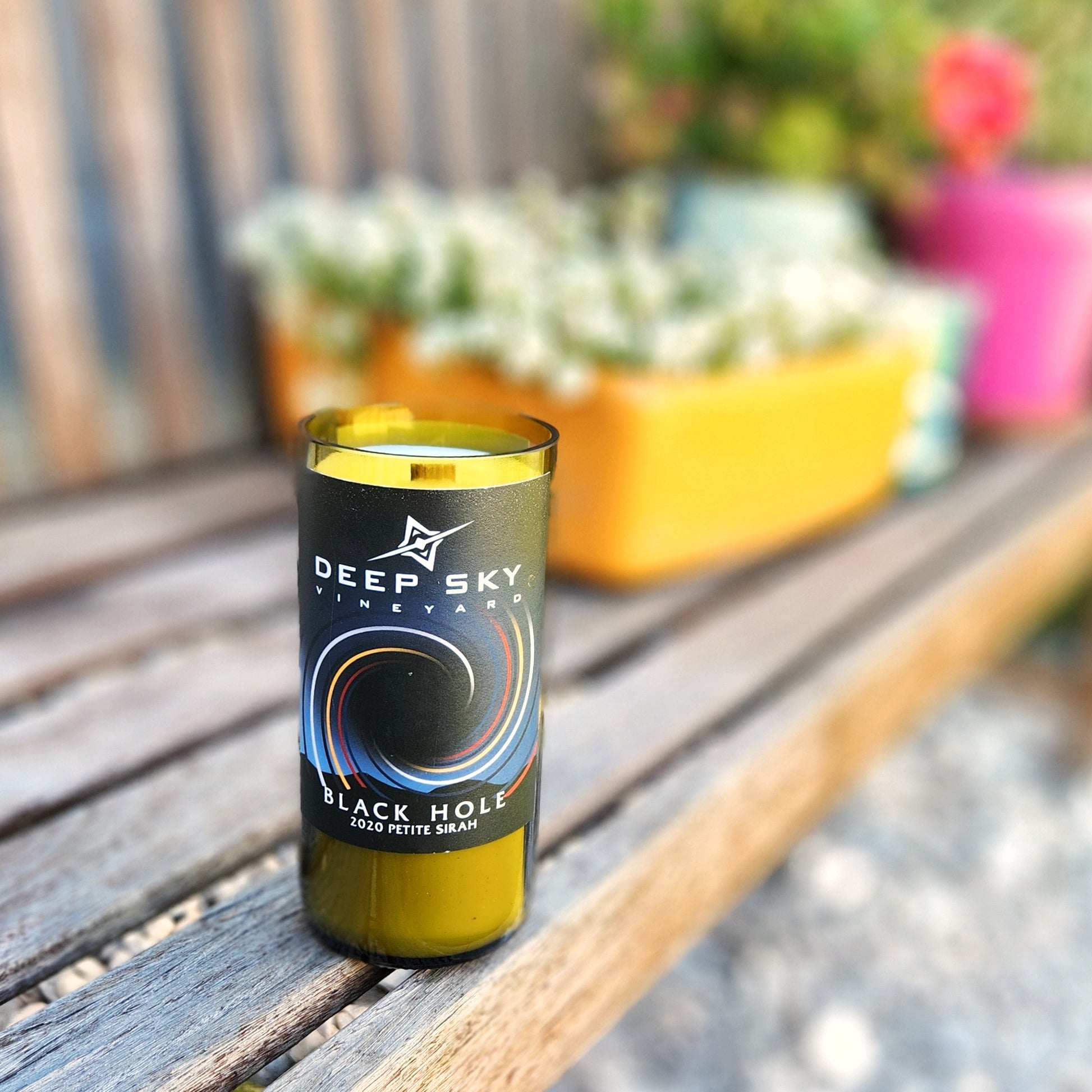 
A recycled glass candle crafted from a green wine bottle, featuring a sleek black label from Deep Sky Vineyard. The label displays a swirling, cosmic-inspired design with the text "Black Hole 2020 Petite Sirah." The candle is placed on a wooden bench outdoors, with vibrant planters filled with greenery and flowers in the blurred background, creating a fresh and inviting atmosphere. The wooden wick and clean finish of the glass rim add a touch of artisanal elegance.